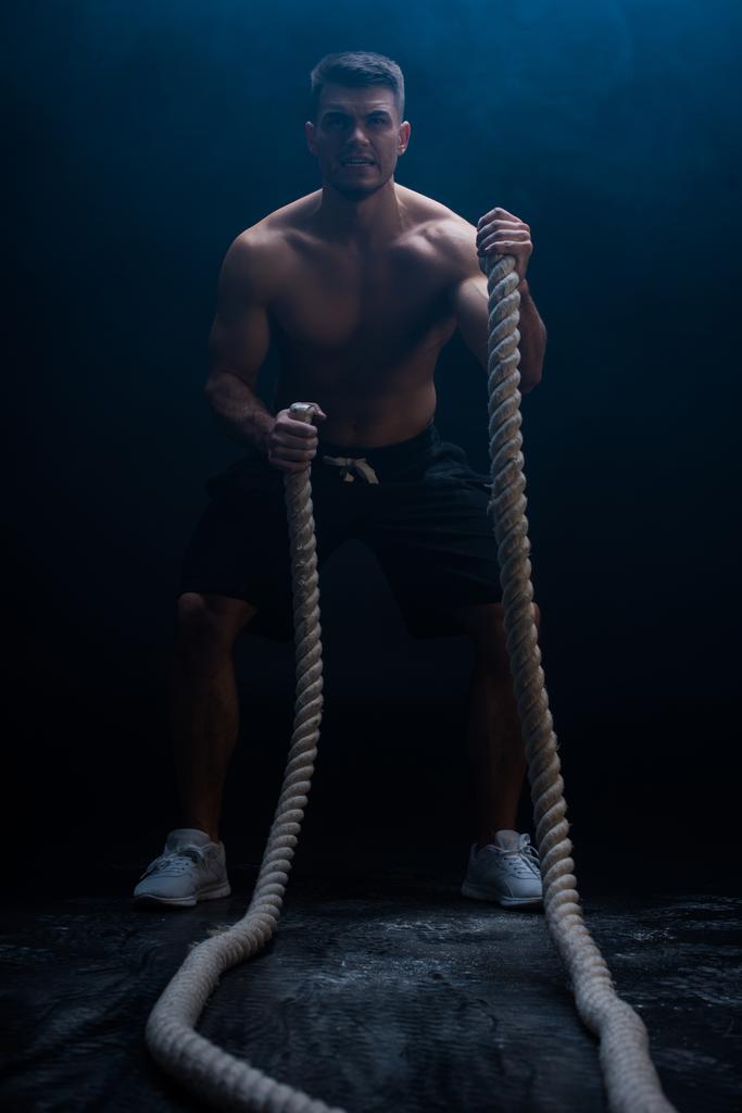 selective focus of sexy muscular bodybuilder with bare torso excising with battle rope on black background with smoke  - Photo, Image