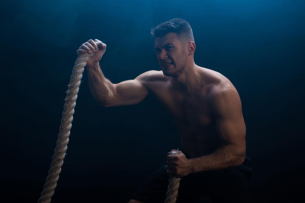angespannter muskulöser Bodybuilder mit nacktem Oberkörper beim Schneiden mit einem Kampfseil auf schwarzem Hintergrund mit Rauch  - Foto, Bild