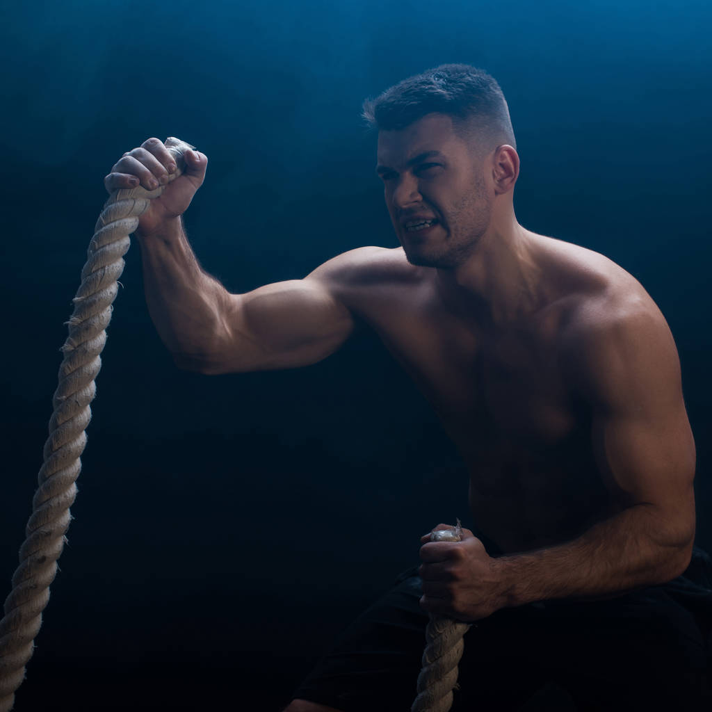 bodybuilder musclé tendu avec torse nu excitant avec corde de combat sur fond noir avec fumée
  - Photo, image