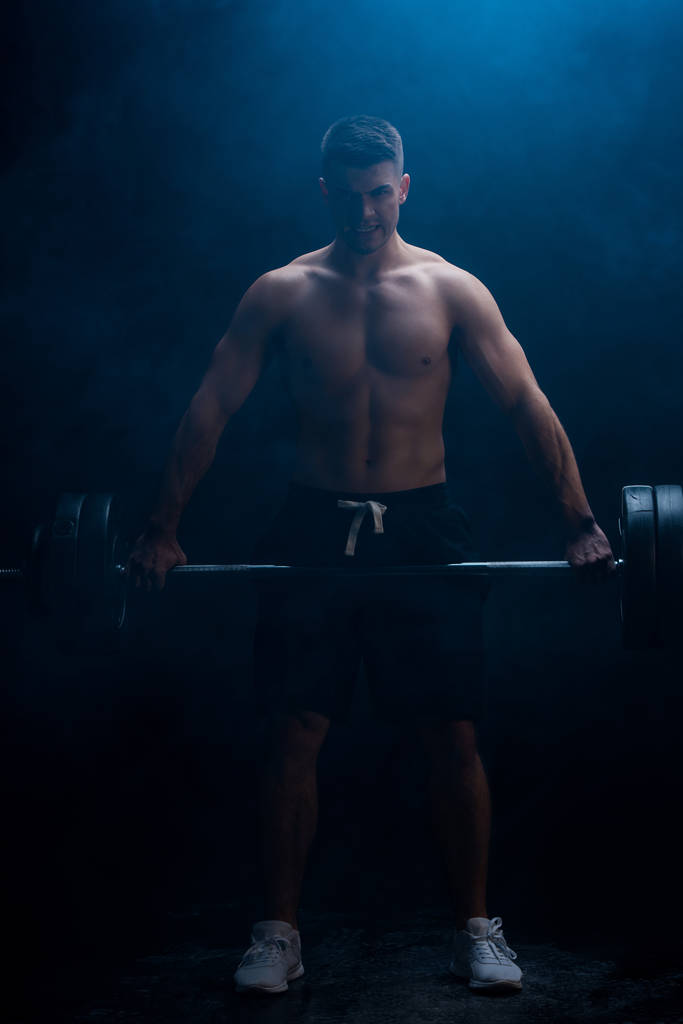 sexy muskulöser Bodybuilder mit nacktem Oberkörper beim Schneiden mit Langhantel auf schwarzem Hintergrund mit Rauch  - Foto, Bild