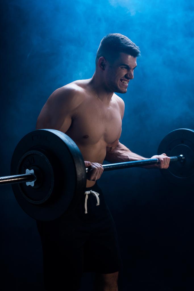 angespannter muskulöser Bodybuilder mit nacktem Oberkörper beim Schneiden mit Langhantel auf schwarzem Hintergrund mit Rauch  - Foto, Bild