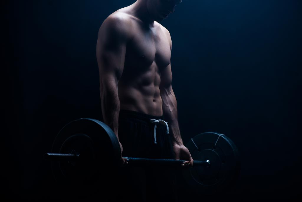 Ausgeschnittene Ansicht eines muskulösen Bodybuilders mit nacktem Oberkörper beim Schneiden mit Langhantel auf schwarzem Hintergrund - Foto, Bild