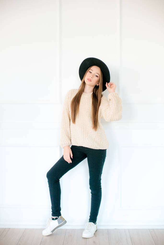Niña adolescente con el pelo largo en un suéter de punto cálido y sombrero de fieltro negro en el estudio sobre un fondo blanco. Moda adolescente
 - Foto, Imagen