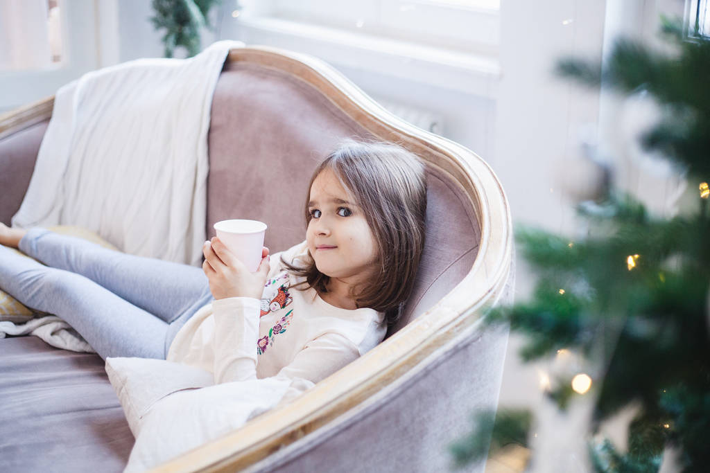 The girl is sitting on the couch with a mug and drinking a drink, the room is decorated for the new year and Christmas - Photo, Image