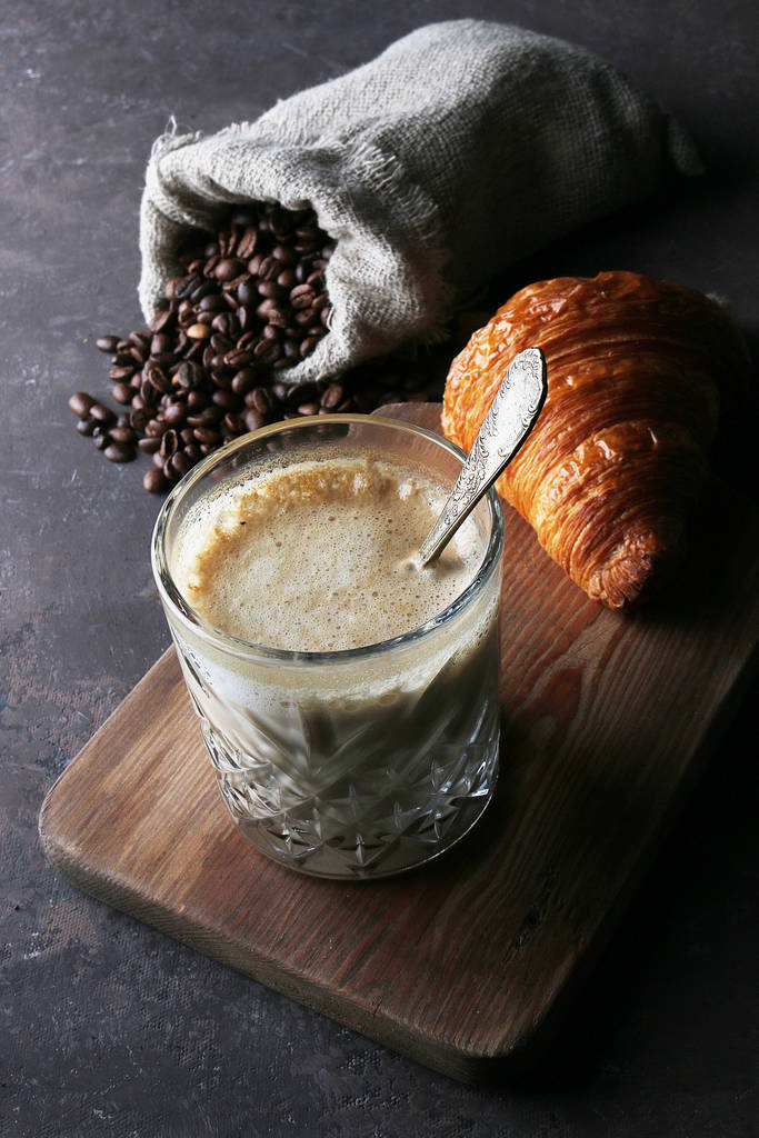 Ποτήρι με latte, κρουασάν και κόκκους καφέ σε τσάντα με λινό σε σκούρο φόντο - Φωτογραφία, εικόνα