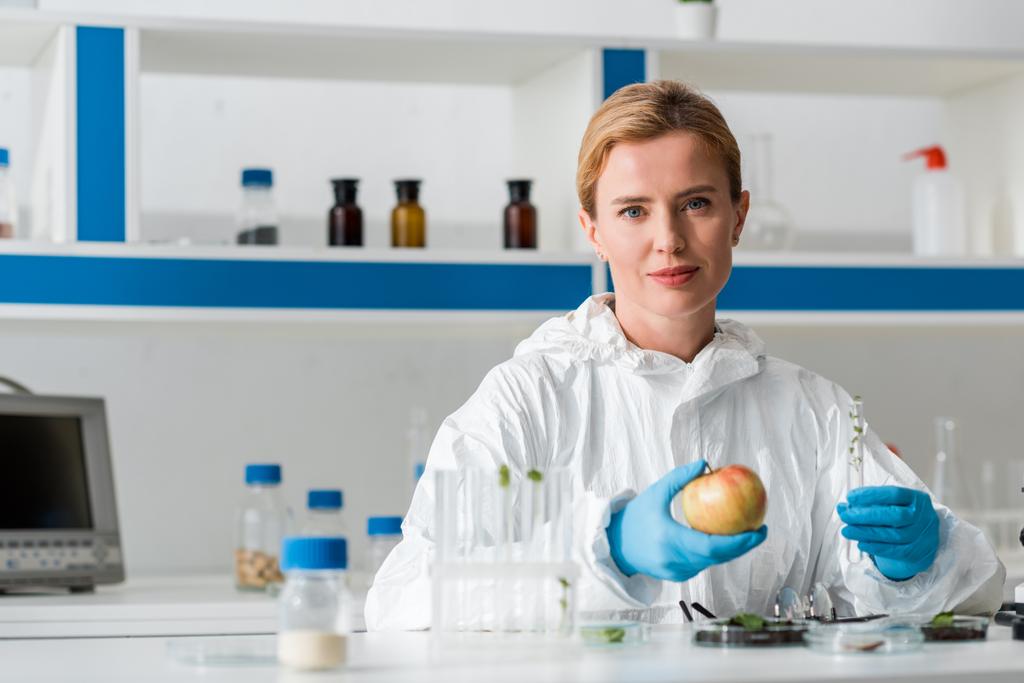 bioloog kijkt naar camera en houdt hele appel en reageerbuis in het lab  - Foto, afbeelding