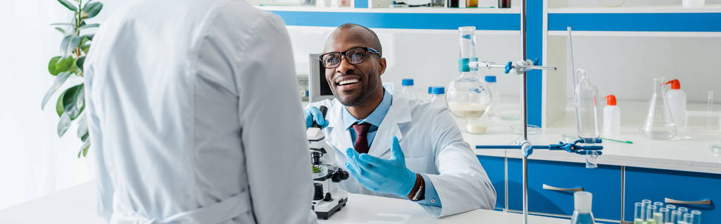 panoramiczne ujęcie uśmiechniętego afrykańskiego biologa patrzącego na swojego kolegę  - Zdjęcie, obraz