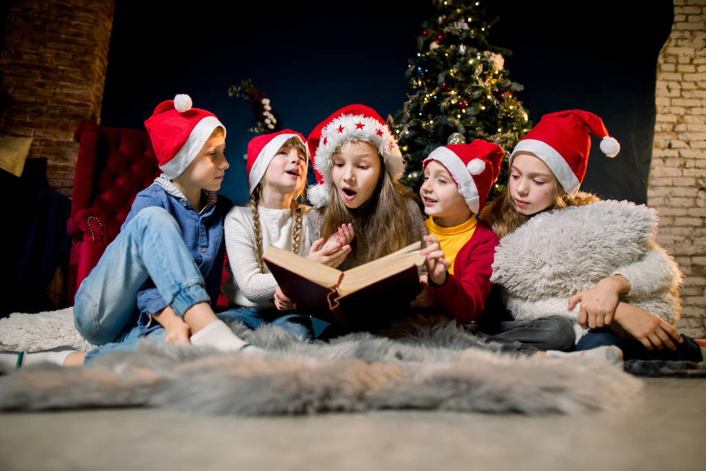 Niños encantadores y hermosos en sombreros de Santas están leyendo cuentos de Navidad y riendo, en el fondo de un árbol de Navidad
. - Foto, Imagen