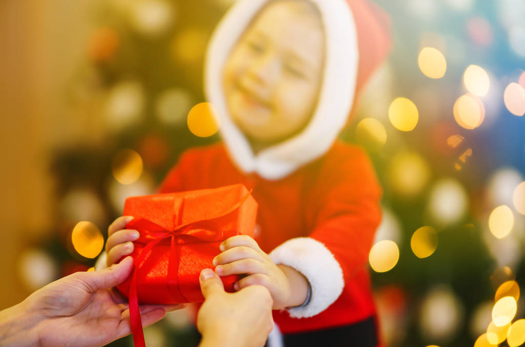 Hands of parent giving a hristmas gift to child. Happy child holding red gift box on the background lights. Kid in santa costume holds christmas present in hands. Holidays, presents, New year concept - Photo, Image