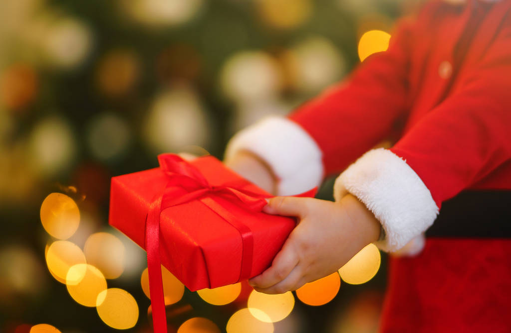 Різдвяний подарунок в руках дитини. Руки маленького хлопчика в костюмі Санти з різдвяним подарунком. Усміхнений смішний хлопчик тримає червону подарункову коробку на тлі світла. Свята, новорічна концепція
. - Фото, зображення