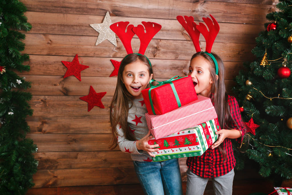 Две счастливые смешные сестрички с подарками возле елки
 - Фото, изображение
