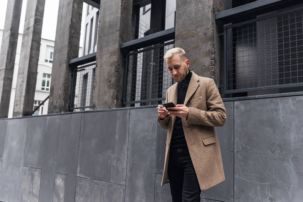 Бизнесмен смотрит на смартфон, стоя в бежевом пальто возле здания
. - Фото, изображение