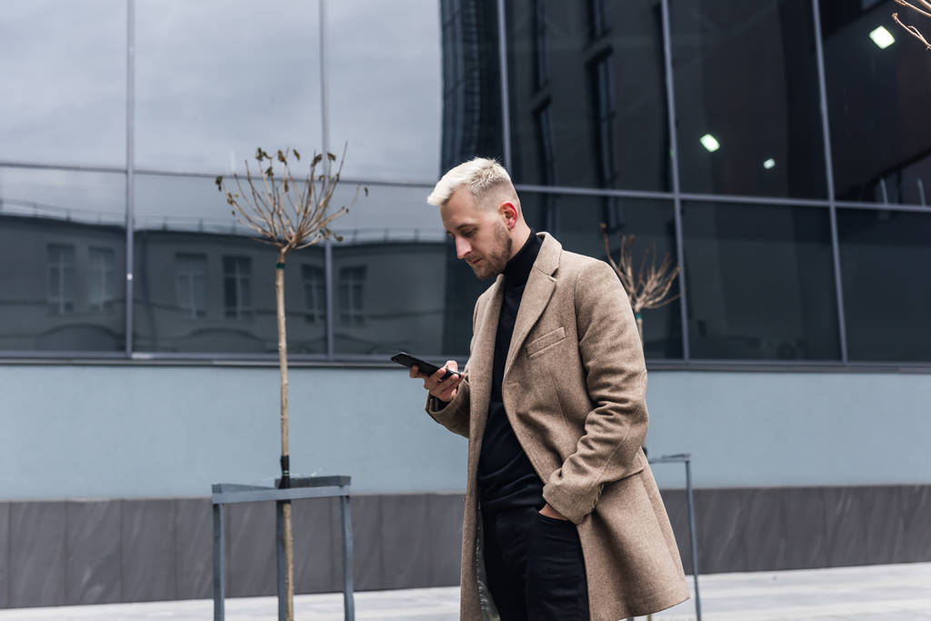 Hombre de negocios mirando el teléfono inteligente mientras está de pie en abrigo beige cerca del edificio
. - Foto, Imagen