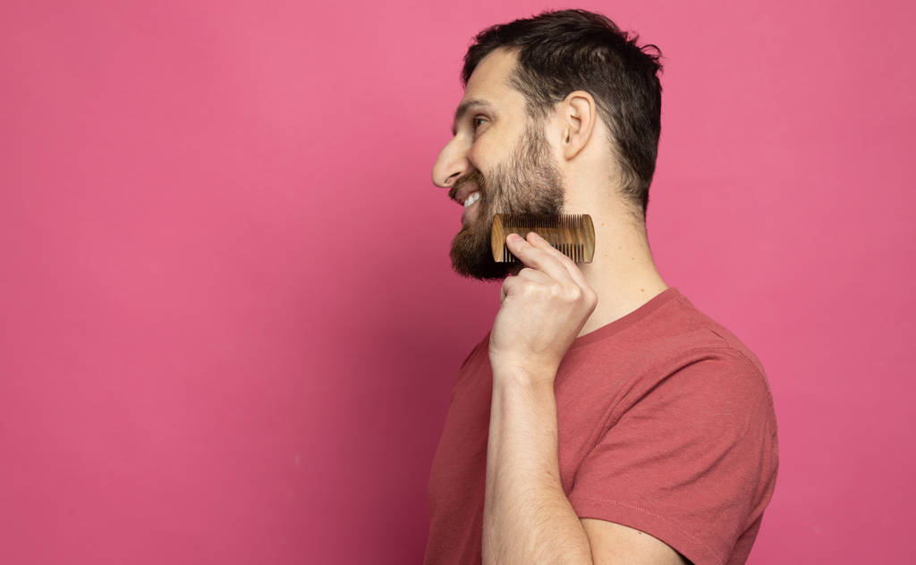 Young bearded man comb his beard and moustache on pink background - Fotoğraf, Görsel