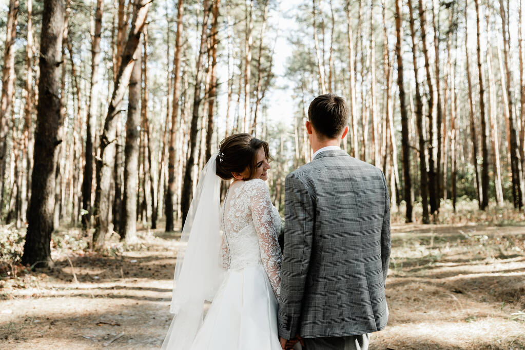 Vonzó pár, akik az esküvőjüket ünneplik az erdőben. Egy portré fiatal boldog vőlegényről és menyasszonyról menyasszonyi ruhában, amint együtt állnak, fogják egymás kezét és egymásra néznek. - Fotó, kép