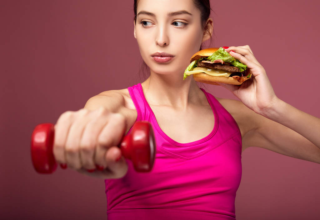 Αθλητικό κορίτσι με ένα όμορφο σώμα κρατά ένα burger και αλτήρες. - Φωτογραφία, εικόνα