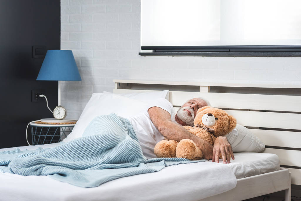 Старий з білим волоссям і бородою спить наодинці з плюшевим ведмедем у своїй спальні. Концепція ночі самотнього чоловіка пенсіонера
 - Фото, зображення