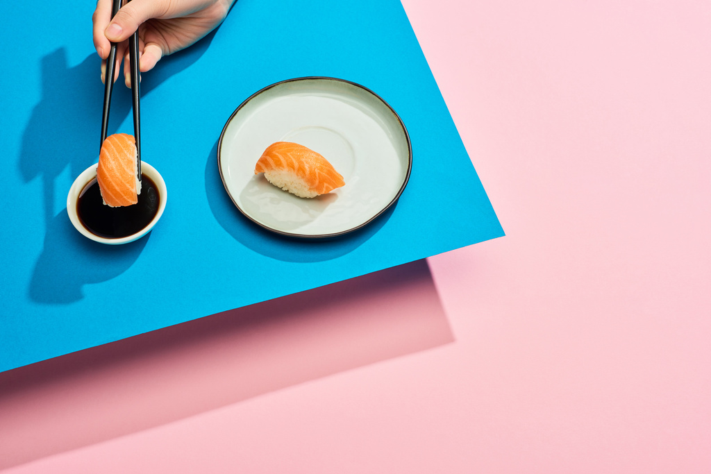 cropped view of woman putting fresh nigiri with salmon into soy sauce on blue, pink background - 写真・画像