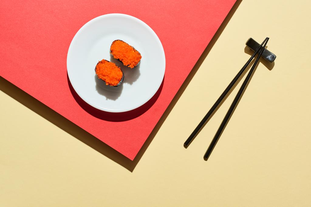 вид сверху на свежий нигири с красной икрой возле палочек для еды на красной и бежевой поверхности
 - Фото, изображение