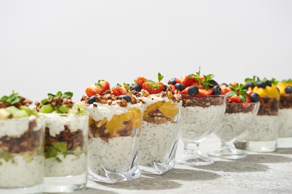 enfoque selectivo de deliciosa granola en vasos con frutas y bayas en superficie de hormigón gris aislado en blanco
 - Foto, Imagen