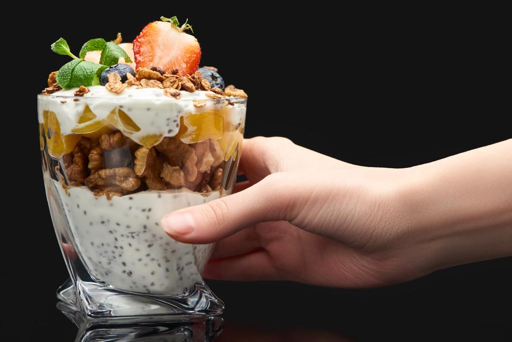 cropped view of woman holding glass with fresh granola with canned peach, berries, walnuts and chia seeds isolated on black - Photo, Image