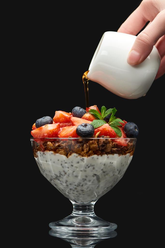 cropped view of woman adding syrup to fresh granola with berries and chia seeds isolated on black - Photo, Image