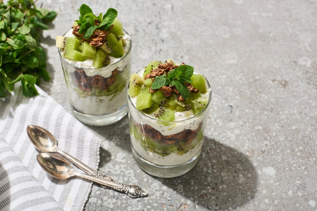 granola fresca con kiwi y yogur en superficie de hormigón gris con servilleta a rayas, cucharas y menta
 - Foto, Imagen