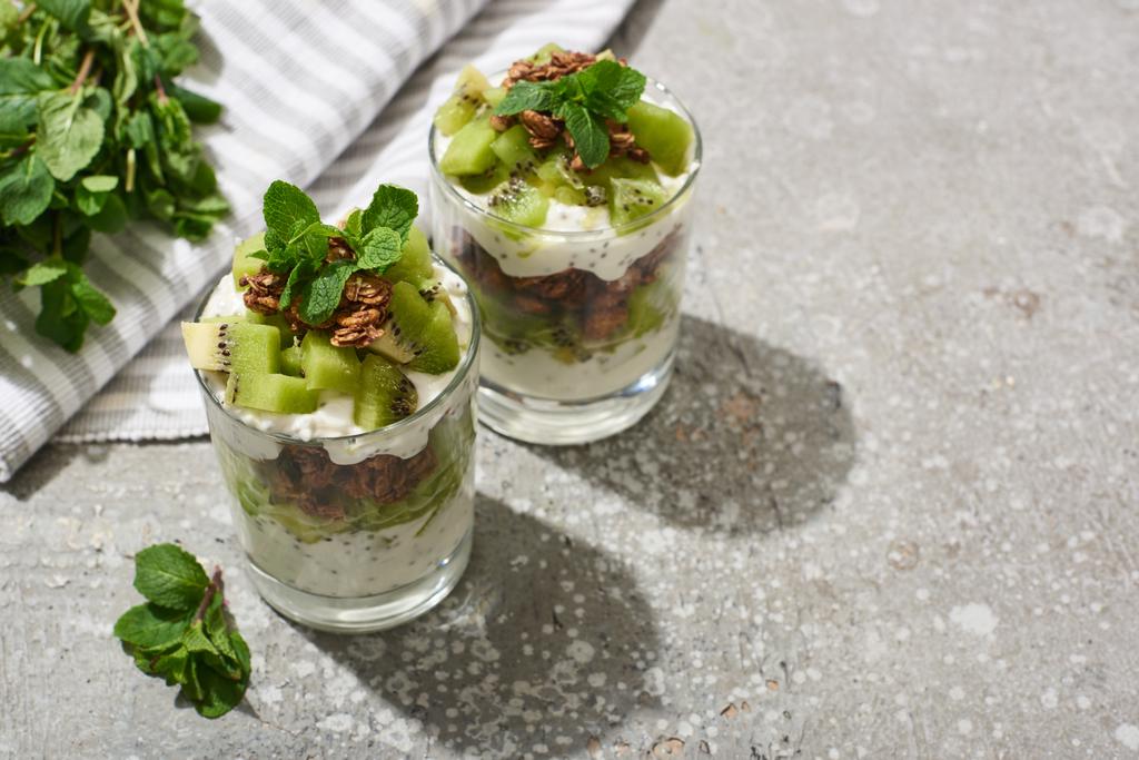 granola fresca con kiwi y yogur en superficie de hormigón gris con servilleta rayada y menta
 - Foto, imagen
