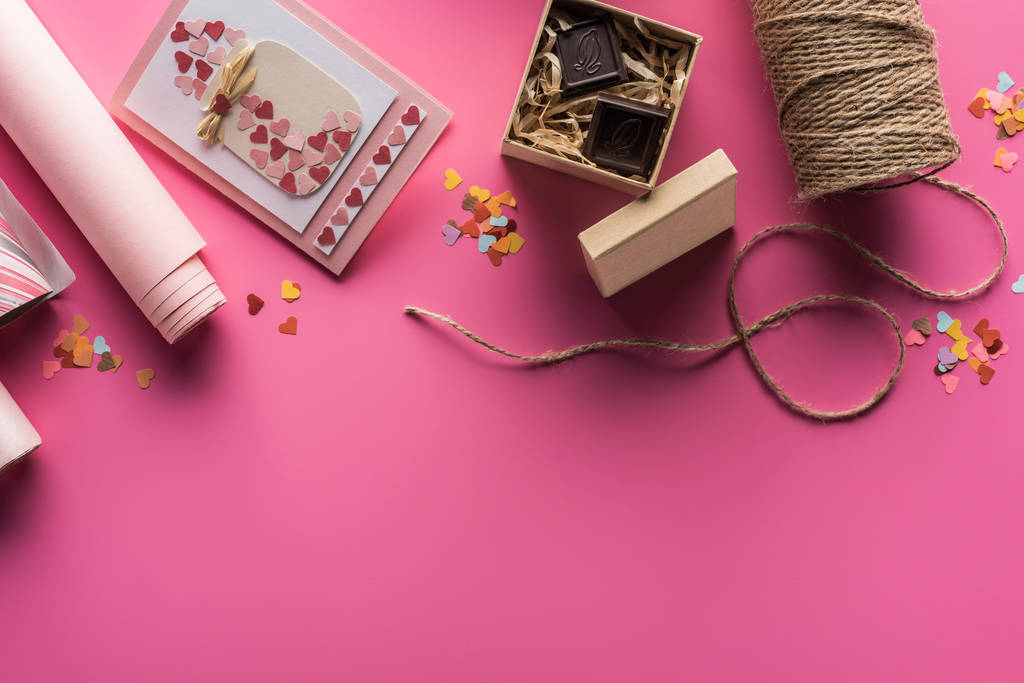 верхний вид украшения валентинки, оберточная бумага, бечевка, подарочная коробка с шоколадом, поздравительная открытка на розовом фоне
 - Фото, изображение