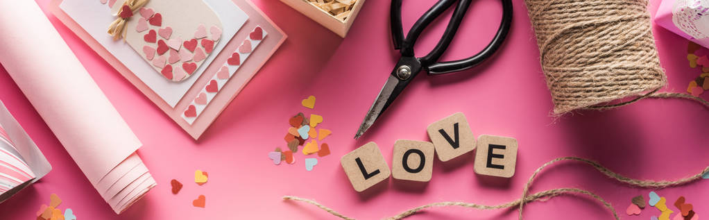 top view of valentines decoration, scissors, wrapping paper, twine, gift, greeting card and love lettering on wooden cubes on pink background, panoramic shot - Photo, Image