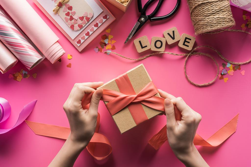 částečný pohled na ženu zdobení dárkové krabice s stuhou v blízkosti valentines ruční práce dodávky na růžovém pozadí - Fotografie, Obrázek