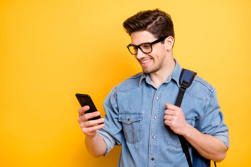 Photo de joyeux beau homme attrayant naviguant à travers le téléphone souriant avec sac à dos derrière fond de couleur vive isolée en jeans denim
 - Photo, image