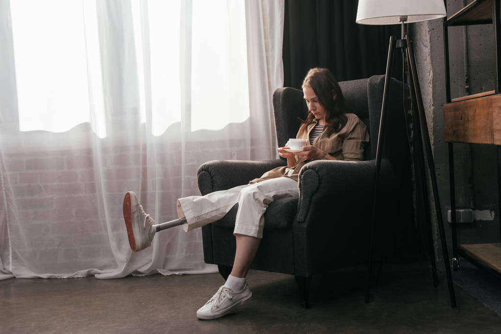junge Frau mit Beinprothese trinkt zu Hause Kaffee im Sessel - Foto, Bild