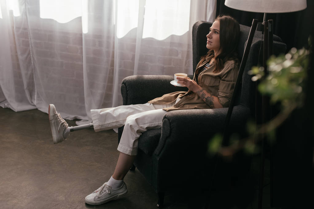Seitenansicht einer jungen Frau mit Beinprothese im Sessel mit Kaffeetasse  - Foto, Bild