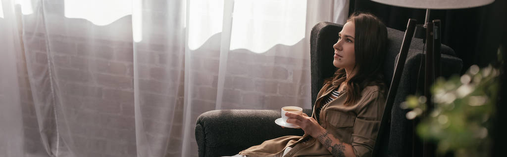 Panoramisch schot van een mooie vrouw met koffiebeker terwijl ze thuis in een fauteuil zit - Foto, afbeelding