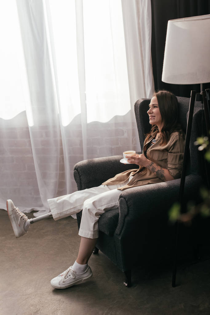 Vista lateral da bela mulher sorridente com prótese perna segurando café enquanto sentado em poltrona
 - Foto, Imagem