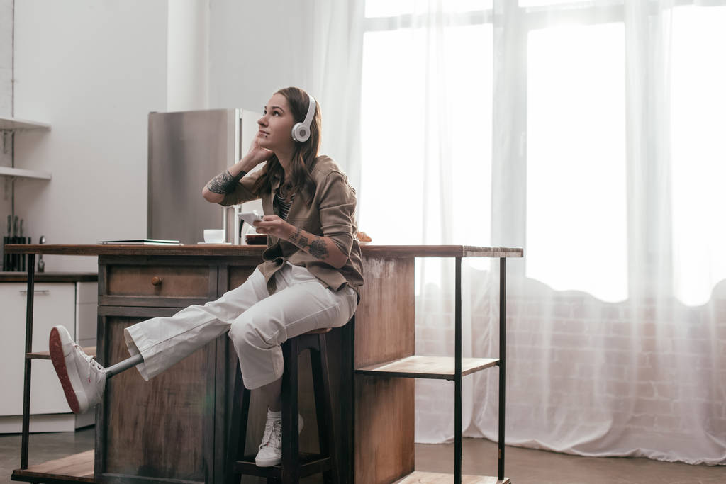 Όμορφη νεαρή γυναίκα με προσθετικό πόδι χρησιμοποιώντας ακουστικά και smartphone στην κουζίνα - Φωτογραφία, εικόνα
