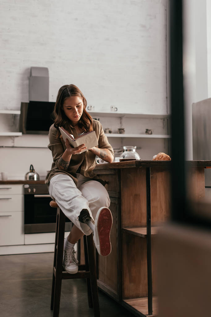 Mulher bonita com prótese perna leitura livro por mesa de cozinha
 - Foto, Imagem