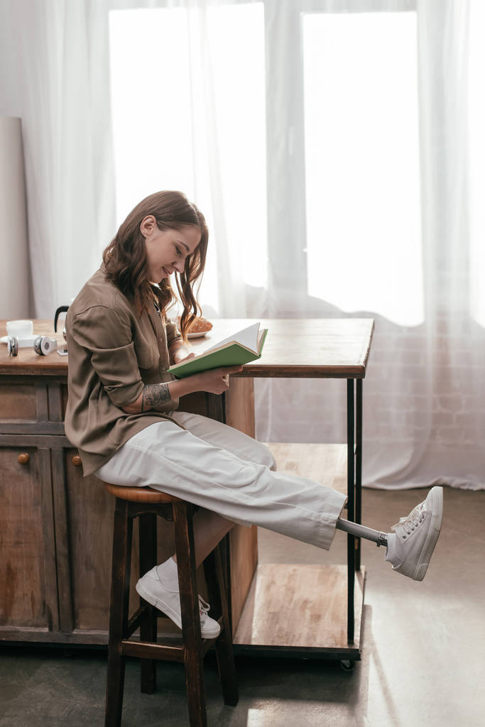 Vista lateral de mujer sonriente con prótesis de pierna libro de lectura al lado de la mesa en casa
 - Foto, imagen
