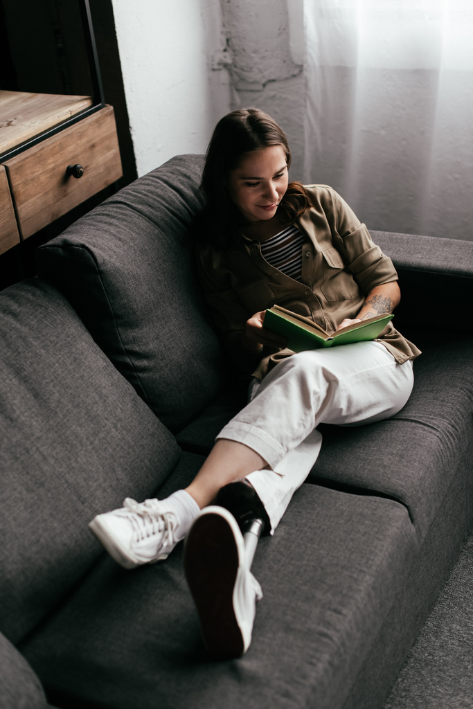 Hochwinkelaufnahme einer lächelnden Frau mit Beinprothese beim Lesen eines Buches auf dem heimischen Sofa - Foto, Bild