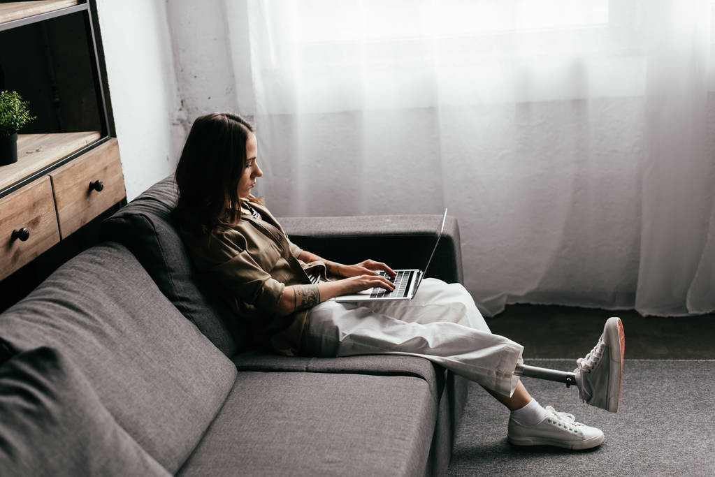 Вид сбоку женщины с протезом ноги, использующей ноутбук на диване в гостиной
 - Фото, изображение