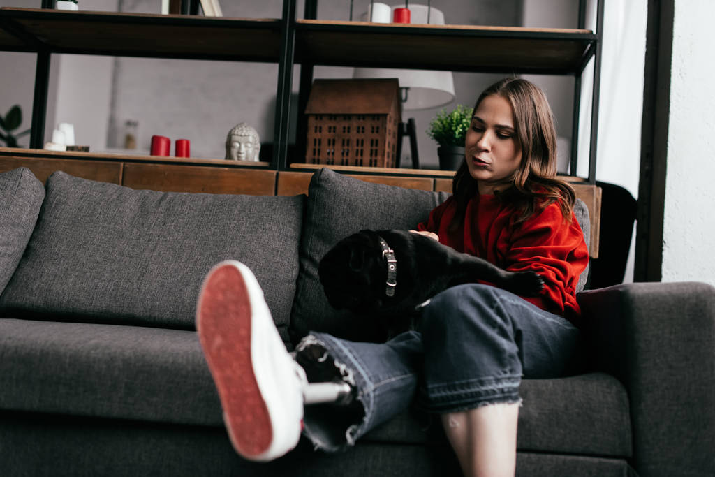 Junge Frau mit Beinprothese streichelt Mops auf Sofa im Wohnzimmer - Foto, Bild