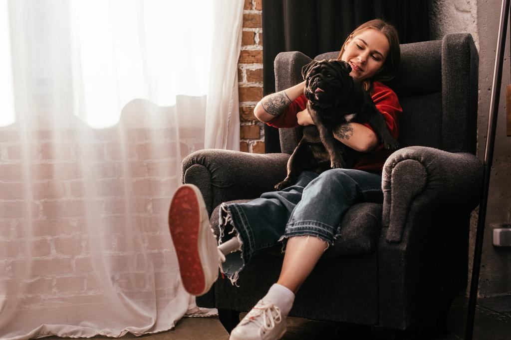 Smiling young woman with leg prosthesis playing with pug in armchair at home - Photo, Image