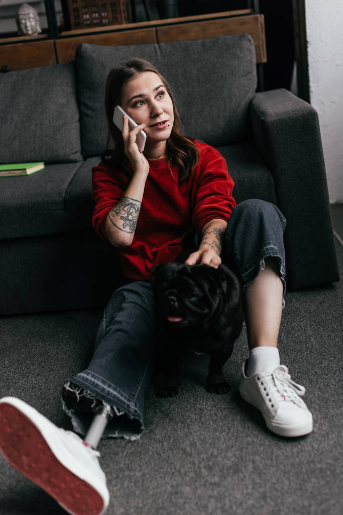 Женщина с протезной ногой разговаривает по смартфону, гладя мопса на полу в гостиной
 - Фото, изображение