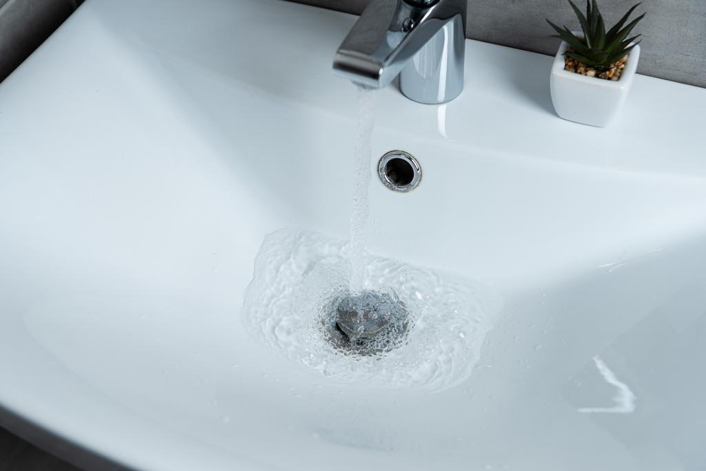 Vysoký úhel pohledu na vodu tekoucí z umyvadla do čistého umyvadla s rostlinou v umývárně - Fotografie, Obrázek