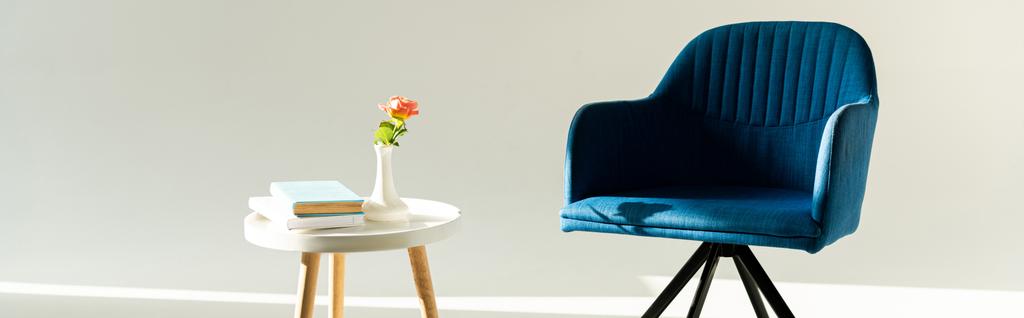 Vazoda çiçekli kahve masasının yanındaki mavi koltuğun panoramik görüntüsü ve gri arka planda kitaplar. - Fotoğraf, Görsel