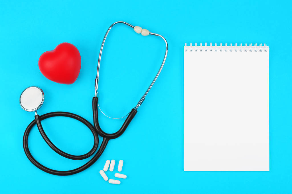 Stethoskop mit blankem Notizblock, rotem Herz und weißen Kapseln auf blauem Hintergrund. - Foto, Bild