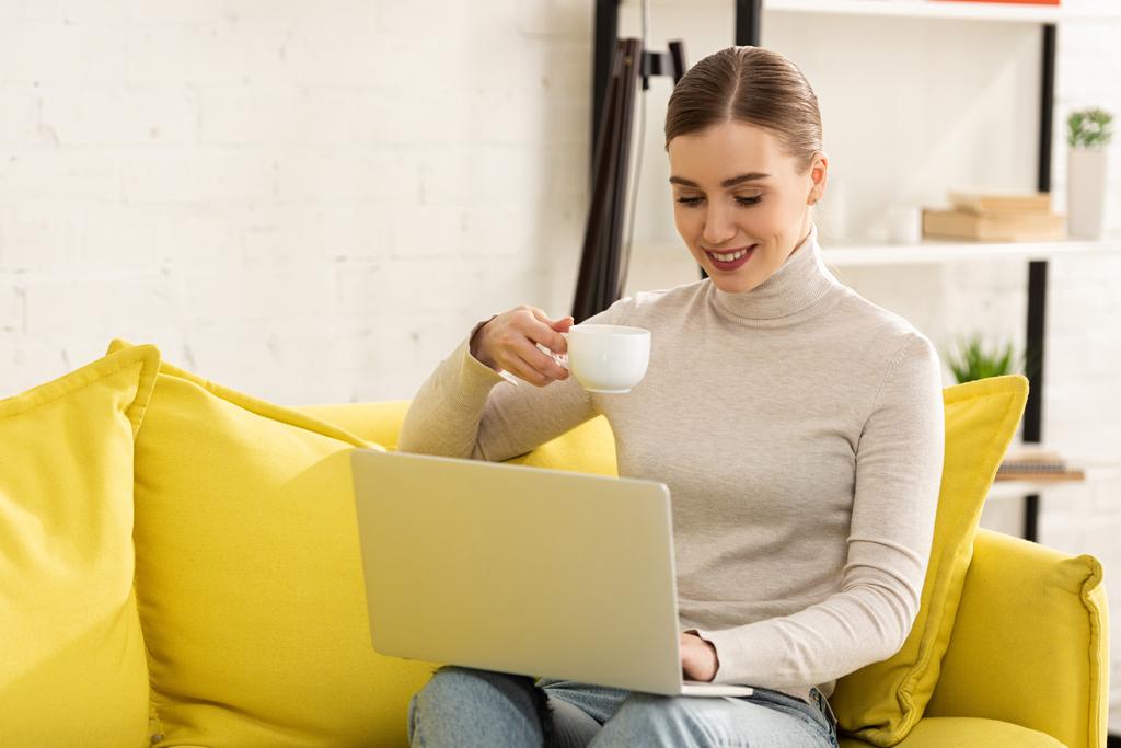 Bella ragazza sorridente bere caffè e lavorare al computer portatile sul divano a casa
 - Foto, immagini