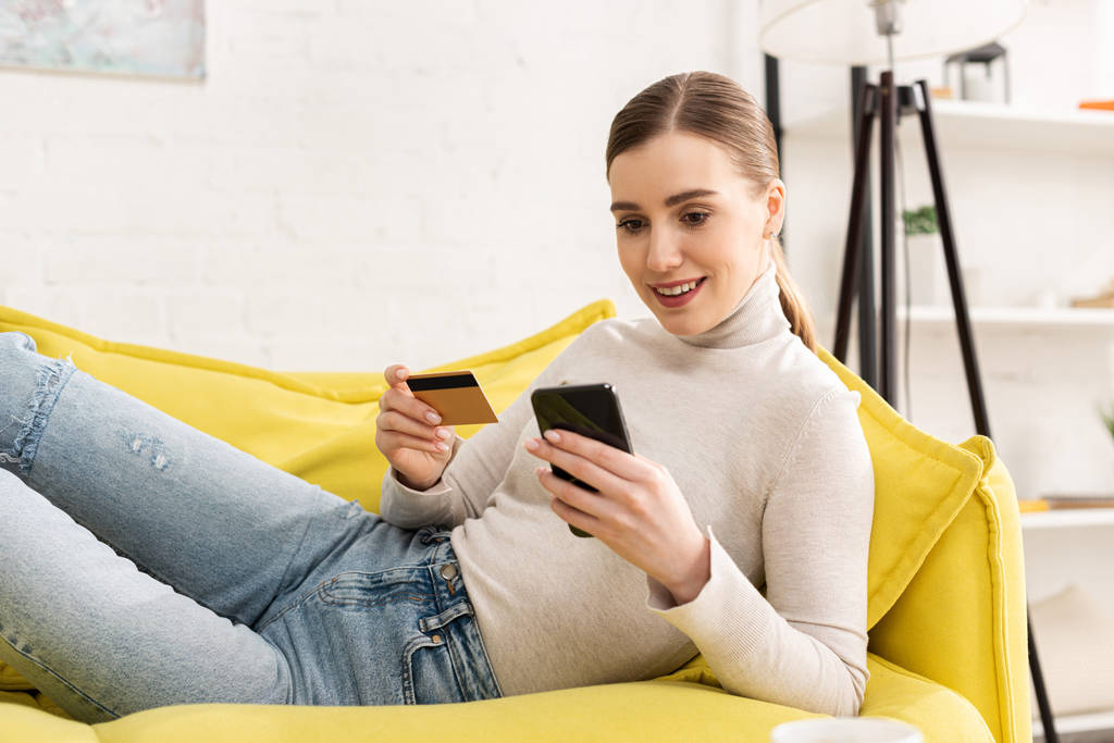Menina sorrindo usando smartphone e segurando cartão de crédito no sofá na sala de estar
 - Foto, Imagem