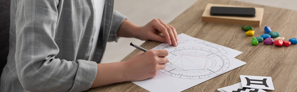 Vista recortada do astrólogo desenhando carta natal por cartas com signos do zodíaco na mesa, tiro panorâmico
 - Foto, Imagem